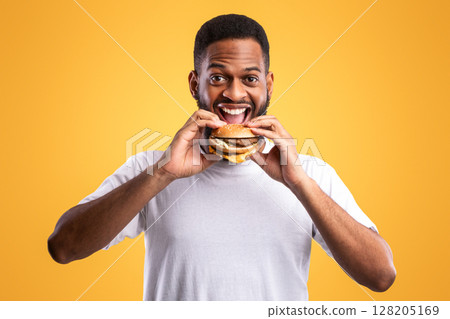 Joyful Hungry African Guy Eating Burger Smiling To Camera Posing Standing On Yellow Background. Studio Shot. Junk Food Eater Enjoying Cheeseburger. Fastfood And Cheat Meal Joyful Hungry African Guy Eating Burger Smiling To Camera Posing Standing On Yellow Background. Studio Shot. Junk Food Eater Enjoying Cheeseburger. Fastfood And Cheat Meal 128205169