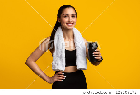 Portrait of sporty beautiful smiling woman in black sportswear and white towel on neck holding shaker with healthy drink, whey protein, fresh water or coffee isolated over orange studio background Portrait of sporty beautiful smiling woman in black sportswear and white towel on neck holding shaker with healthy drink, whey protein, fresh water or coffee isolated over orange studio background 128205172