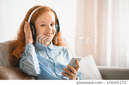 Favorite Song. Smiling Red-Haired Girl Sitting On The Couch At Home, Listening To Music And Touching Wireless Headphones, Holding Her Mobile Phone And Looking At Camera, Enjoying Free Time 128205175