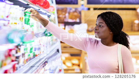 Beautiful black woman buying products at dairy department of huge supermarket. Young African American female consumer shopping at mall, choosing quality food products on sale 128205176