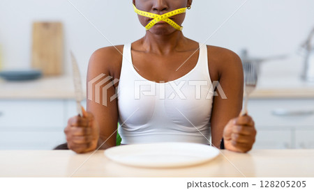 Diet Restrictions. Hungry African American Lady Having Tied Mouth With Measuring Tape Sitting Near Empty Plate, Looking At Camera In Kitchen At Home. Fasting And Weight Loss, Food Craving And Hunger 128205205