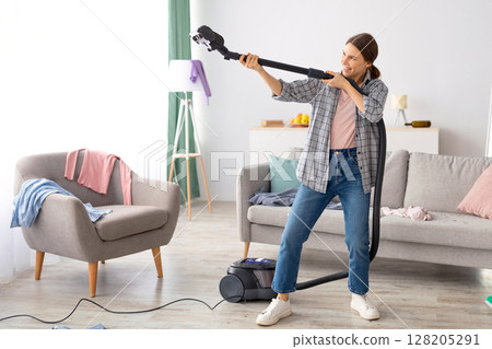 Positive young lady holding vacuum cleaner like rifle, ready to perform cleanup at messy room after party, copy space. Millennial female student at dirty apartment, prepared for hoovering Positive young lady holding vacuum cleaner like rifle, ready to perform cleanup at messy room after party, copy space. Millennial female student at dirty apartment, prepared for hoovering 128205291