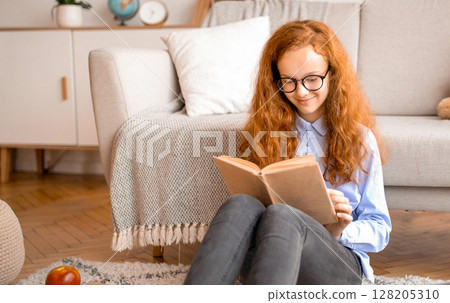 Youth And Literature. Full length of positive gen z girl in eyeglasses holding and reading paper book. Schoolgirl sitting on the floor carpet with laptop at home in cozy bedroom, leaning on couch 128205310
