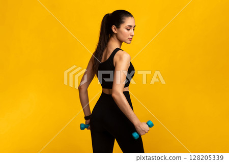 Fitness Model. Portrait Of Confident Lady In Black Sportswear Exercising With Two Dumbbells Posing Isolated Over Orange Background, Turning Back And Looking Down. Studio Shot, Free Space, Banner 128205339