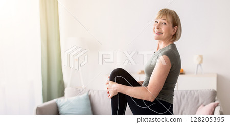 Sporty senior woman stretching her leg and smiling at camera indoors, free space. Attractive mature lady doing flexibility exercises, leading healthy lifestyle. Domestic trainings during coronavirus 128205395