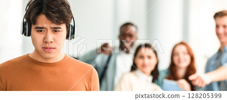 Group Conflict. Sad asian guy in headphones standing away alone, suffering humiliation and public disgrace, crowd of bad people bullying pointing fingers at victim, laughing. Racial Discrimination 128205399