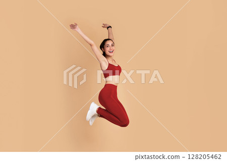 Full Length Portrait Of Happy Excited Smiling Young Woman In Sports Bra And Leggins Jumping Up And Raising Arms Up Isolated On Pastel Background, Celebrating Success, Studio Shot, Copy Space. Full Length Portrait Of Happy Excited Smiling Young Woman In Sports Bra And Leggins Jumping Up And Raising Arms Up Isolated On Pastel Background, Celebrating Success, Studio Shot, Copy Space. 128205462