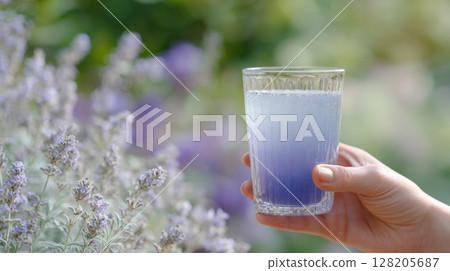 Hand holding a glass of lavender drink surrounded by blooming flowers in a garden setting 128205687