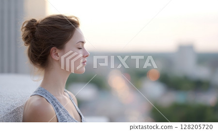 Young woman with closed eyes is enjoying a moment of tranquility on a rooftop, overlooking a blurred cityscape at sunset, embodying peace and relaxation 128205720