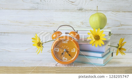 Alarm clock and stack of books with fresh flowers between pages, as bookmarks. On white wooden background, concept back to school, education. Alarm clock and stack of books with fresh flowers between pages, as bookmarks. On white wooden background, concept back to school, education. 128205747