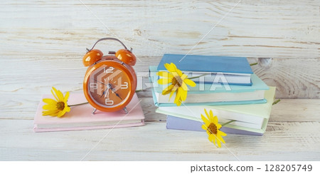 Alarm clock and stack of books with fresh flowers between pages, as bookmarks. On white wooden background, concept back to school, education. 128205749