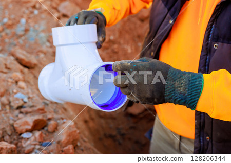Construction worker carefully fits white PVC sewage pipe at trench, on construction site, plumbing work day 128206344