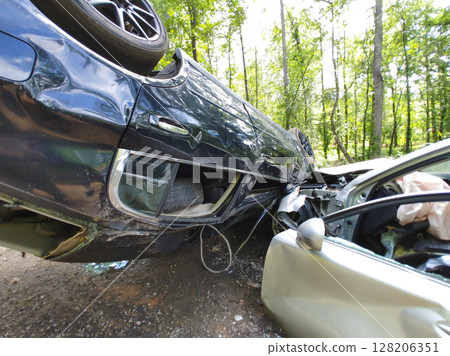 Two damaged cars involved in collision, with one vehicle flipped on its side surrounded by trees. 128206351