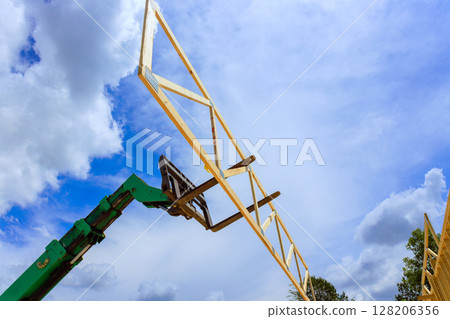 Forklift telehandler machinery raises wooden trusses beams high in air as fluffy clouds fill blue sky during workday. 128206356