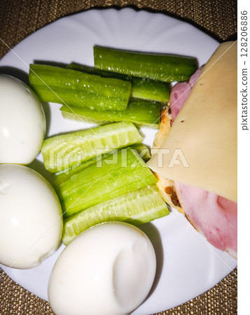 Freshly prepared plate with boiled eggs, crunchy cucumber sticks, and a slice of ham topped with cheese, showcasing a nutritious meal idea for healthy eating concept 128206886