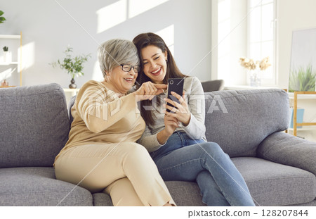 Old mother and adult daughter sit on sofa looking photos on phone, sharing laughter and cherished memories. Old mother and adult daughter love different generations family love of mother and daughter. Old mother and adult daughter sit on sofa looking photos on phone, sharing laughter and cherished memories. Old mother and adult daughter love different generations family love of mother and daughter. 128207844