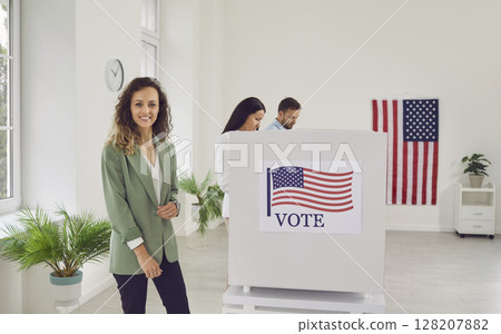 Voter woman looking at camera standing at vote center near voting booth on election day. Voter woman looking at camera standing at vote center near voting booth on election day. 128207882