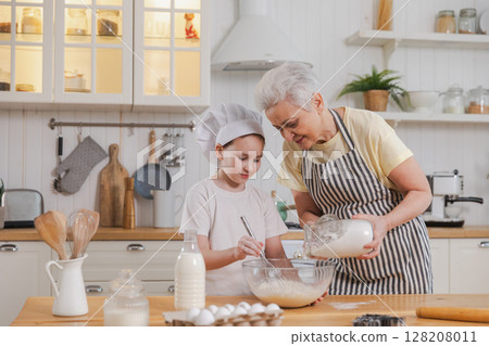 Happy family in kitchen. Grandmother and granddaughter child cook in kitchen together. Grandma teaching kid girl knead dough bake cookies. Household teamwork helping family generations concept 128208011