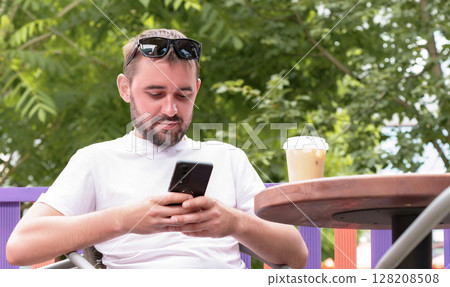 Bearded man wearing sunglasses and a white t shirt, sipping iced coffee at an outdoor cafe, using a smartphone to browse social media while enjoying a sunny summer day Bearded man wearing sunglasses and a white t shirt, sipping iced coffee at an outdoor cafe, using a smartphone to browse social media while enjoying a sunny summer day 128208508