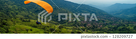 A paraglider with an orange canopy glides over lush green hills and a distant village. Concept of adventure and nature. For travel and outdoor activity promotions. A paraglider with an orange canopy glides over lush green hills and a distant village. Concept of adventure and nature. For travel and outdoor activity promotions. 128209727