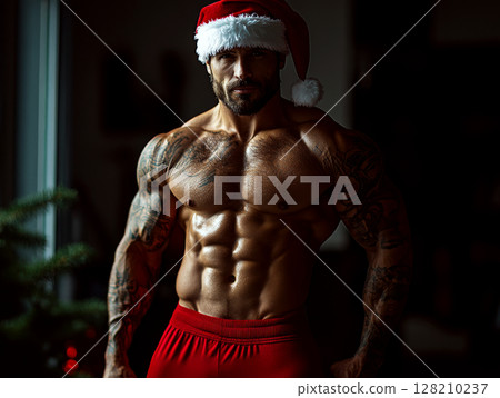 A muscular man wearing red shorts and a Santa hat stands confidently in a dimly lit room. Concept of holiday fitness. For fitness-themed holiday content. 128210237