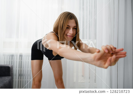 Young woman in black sportswear performing a forward stretch with clasped hands. 128210723