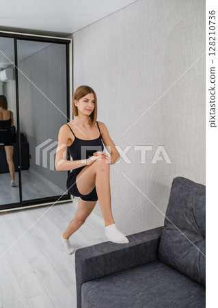 Young woman doing lunge stretch with foot on sofa. Young woman doing lunge stretch with foot on sofa. 128210736