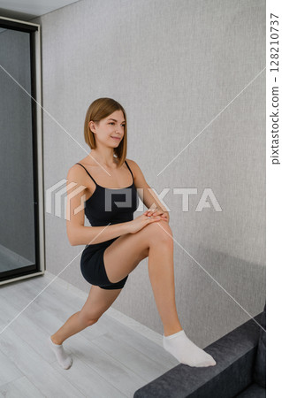 Young woman doing lunge stretch with foot on sofa. Young woman doing lunge stretch with foot on sofa. 128210737