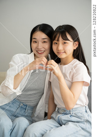 Mother and daughter making a heart shape with their hands indoors 128210822
