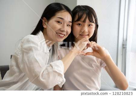 Mother and daughter making a heart shape with their hands indoors 128210823