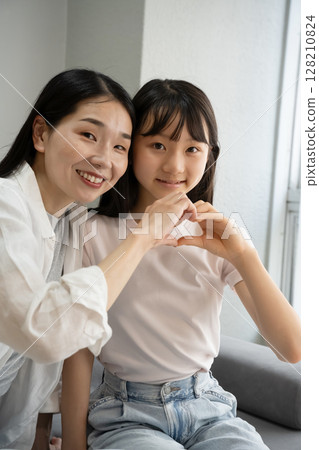 Mother and daughter making a heart shape with their hands indoors 128210824