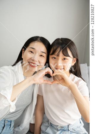 Mother and daughter making a heart shape with their hands indoors 128210827