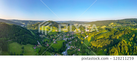 This aerial view of Tanvaldsky Spicak in the Jizera Mountains highlights green hills, dense forests, and charming villages under a clear blue sky in the morning light. This aerial view of Tanvaldsky Spicak in the Jizera Mountains highlights green hills, dense forests, and charming villages under a clear blue sky in the morning light. 128211259