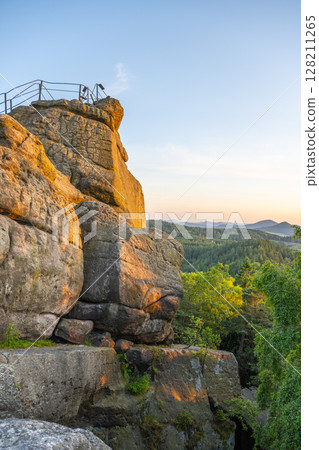 At sunset, the warm light casts a golden glow on the sandstone formations of Popov Cliffs. The lush green landscape provides a stunning backdrop as evening descends over Czechia. 128211265