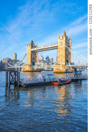 Morning at Tower Bridge features clear skies and sunlight reflecting off the River Thames. Visitors admire the iconic structure while boats glide by, showcasing a vibrant London. 128211268