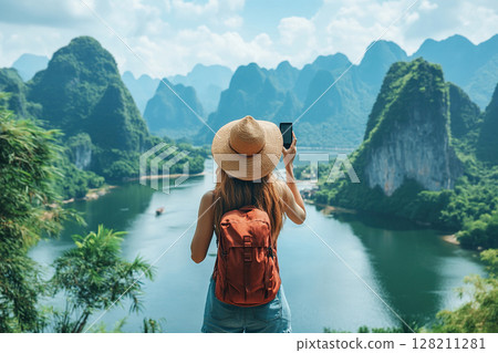 A female traveler in a hat with a backpack on her back takes a photo of an amazing landscape with rocks, sea and blue sky on her smartphone during her vacation. A female tourist during her vacation. 128211281