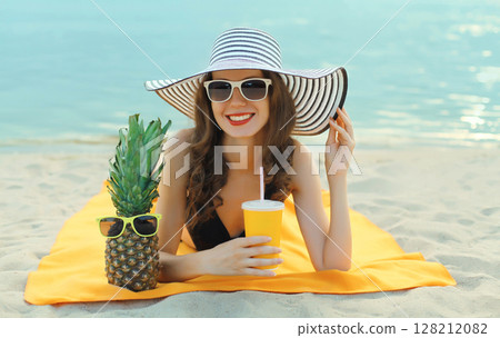 Summer vacation, happy smiling young woman with pineapple in hat drinking juice lying on sea beach 128212082