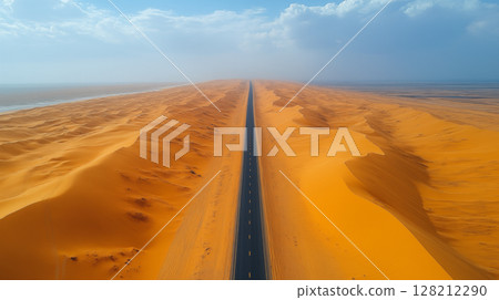 Desert highway extending to horizon through golden sand dunes under blue sky, aerial view of empty road in dramatic arid landscape for travel concept 128212290