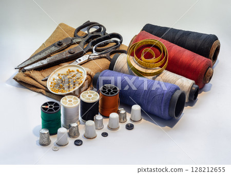 Steel scissors and Pins in small round tray on Sackcloth with Measuring tape on Spools of thread of various colour and Thimbles, Black buttons on white background. 128212655