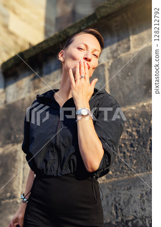 Portrait young adult elegant woman black dress and sunglasses blowing air kiss in golden sunlight warm sunset backlit light against historic cathedral building. Female person walking city street 128212792
