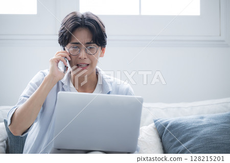 A man wearing glasses apologizing while talking on his smartphone and looking at a computer 128215201