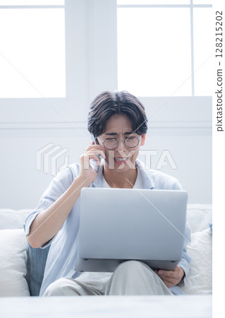 A man wearing glasses apologizing while talking on his smartphone and looking at a computer 128215202