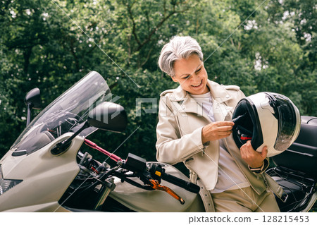 Smiling senior woman with gray hair checking motorcycle helmet, symbol of freedom, active lifestyle Smiling senior woman with gray hair checking motorcycle helmet, symbol of freedom, active lifestyle 128215453