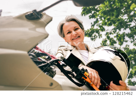 Smiling senior woman with gray hair holding helmet next to motorcycle, confident and free outdoors 128215462