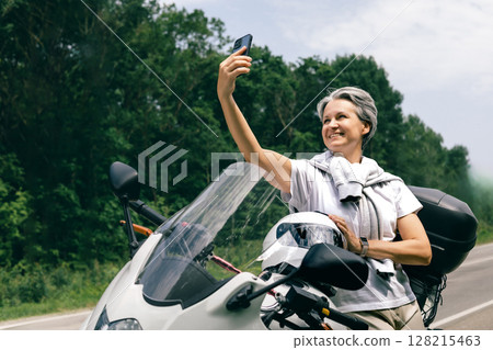 Smiling mature woman with gray hair taking selfie on motorcycle, freedom and joy in later years 128215463