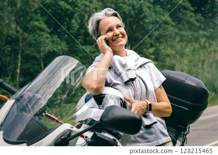 Happy senior woman with gray hair talking on smartphone by her motorbike on a forest country road 128215465