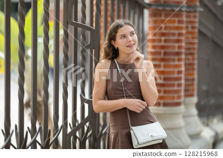 Pretty young woman traveler in summer dress with handbag posing near Hospital de la Santa Creu i Sant Pau in Barcelona on spring day 128215968