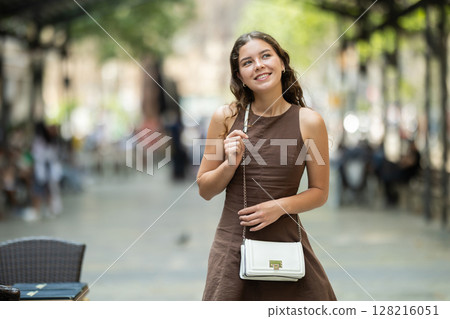 Cute young girl beautifully dressed in modern summer dress walking along city streets and choosing cafe to have snack and have good time Cute young girl beautifully dressed in modern summer dress walking along city streets and choosing cafe to have snack and have good time 128216051