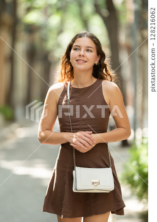 Beautiful smiling young lady wearing short dress walking shady tree lined street and relax in summer weather with sunshine outdoor 128216062