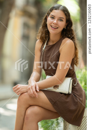 Beautiful young lady wearing dress walking shady street and sitting concrete flower bed with plants, taking break from heat of day 128216063